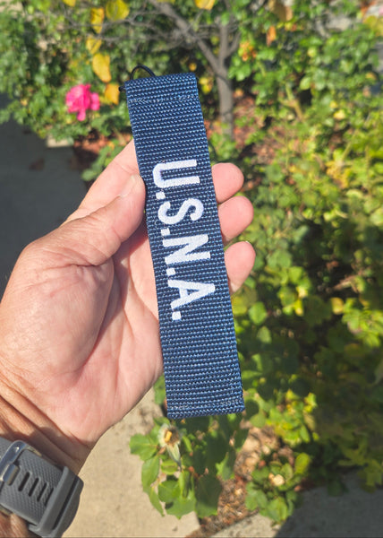 Hand holding a blue woven patch with 'U.S.N.A.' text against a blurred natural background