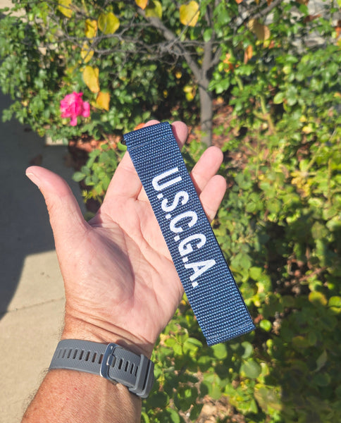 Hand holding a blue U.S.C.G.A. luggage tag with greenery in the background